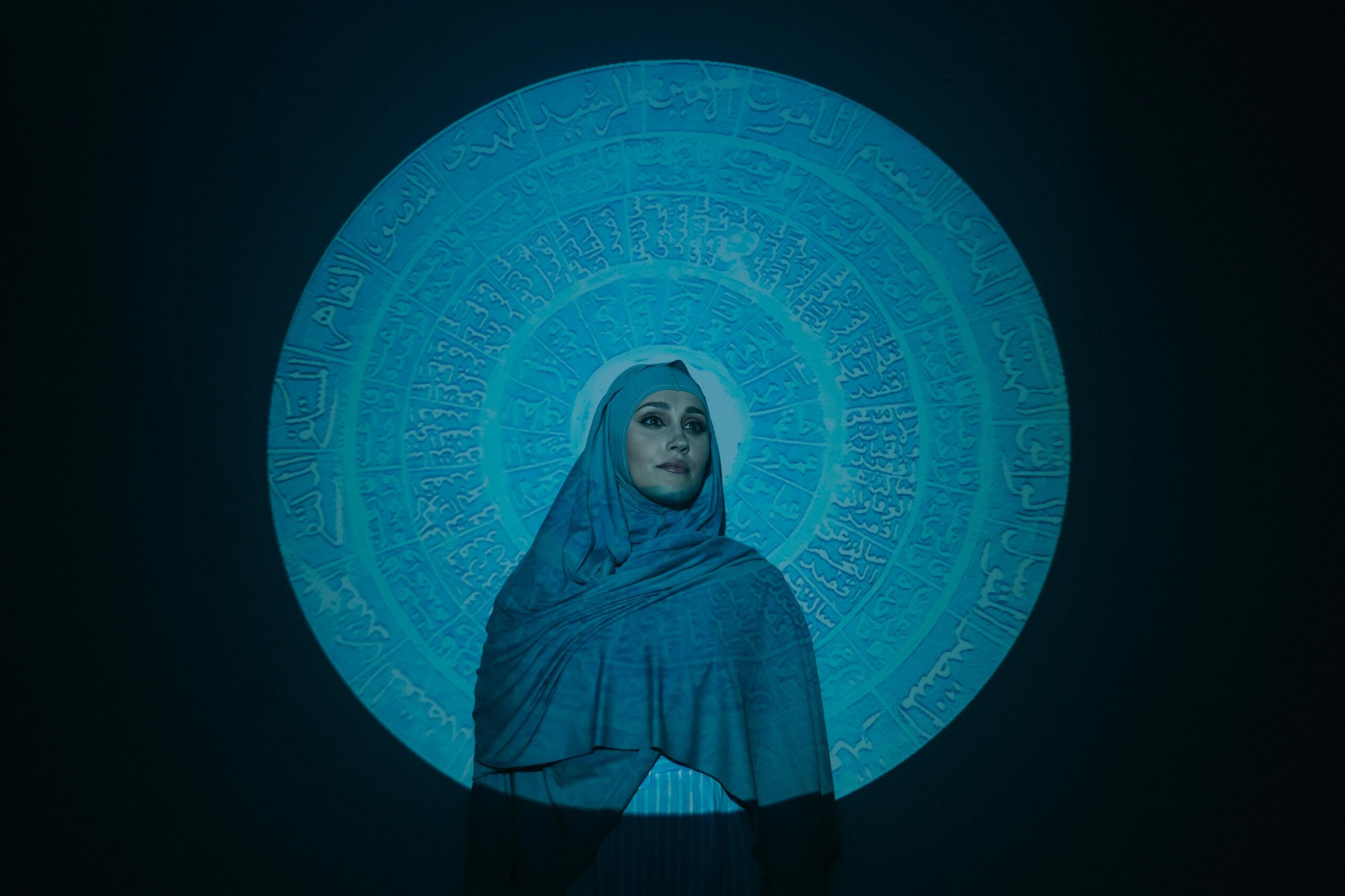 A woman in a hijab is illuminated by a circular Arabic calligraphy projection.